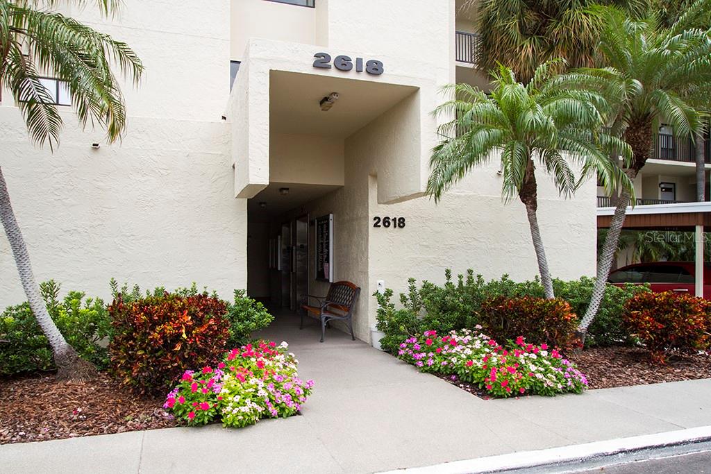 The exterior entrance of a multi-story building features a covered walkway, landscaping, and palm trees — 2618 COVE CAY DRIVE UNIT 109, CLEARWATER, FL