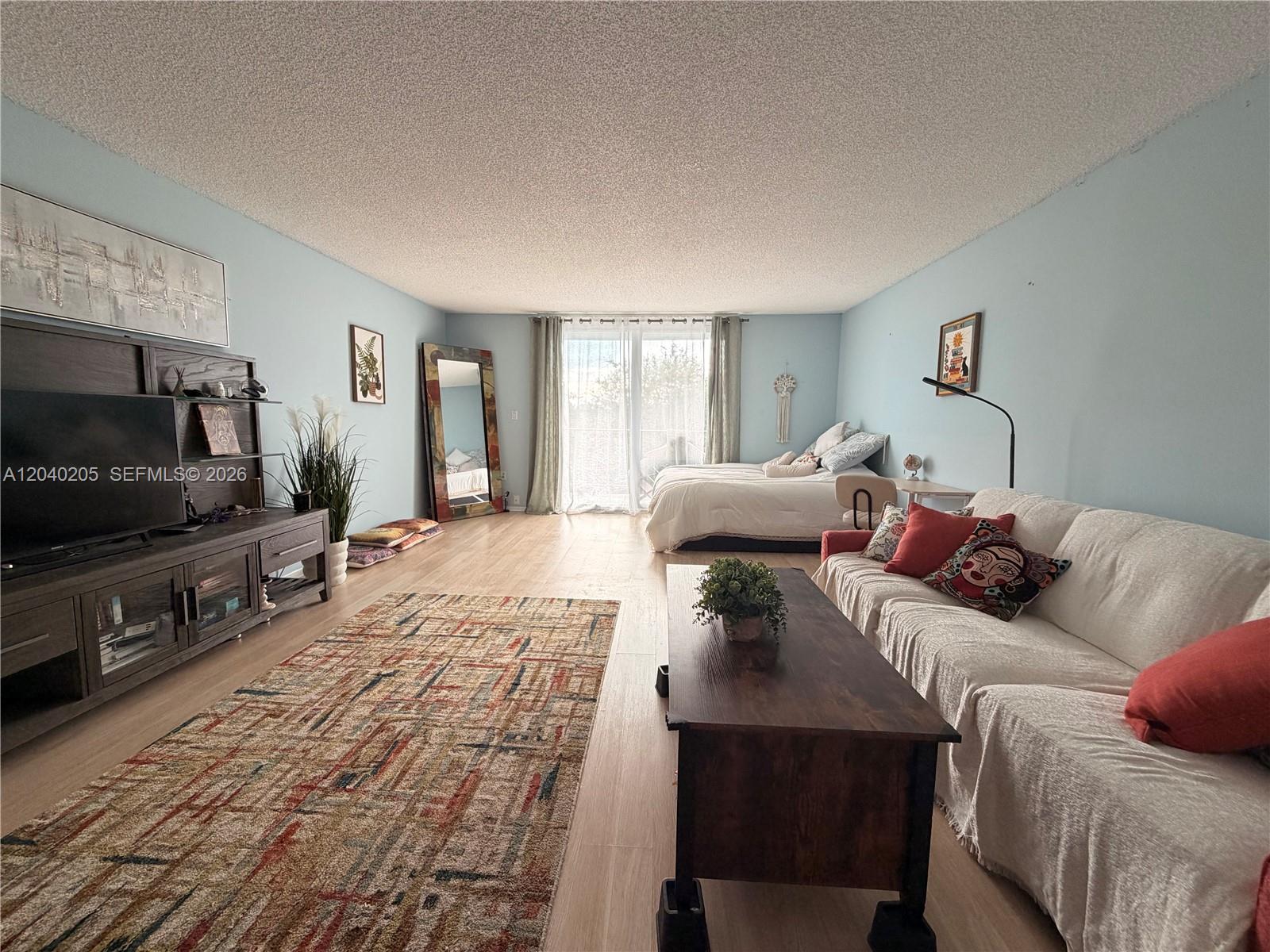 A studio apartment features light blue walls, wood-look flooring, a living area, and a bed — 12500 NE 15th Ave # 312, North Miami FL 33161, NORTH MIAMI, FL