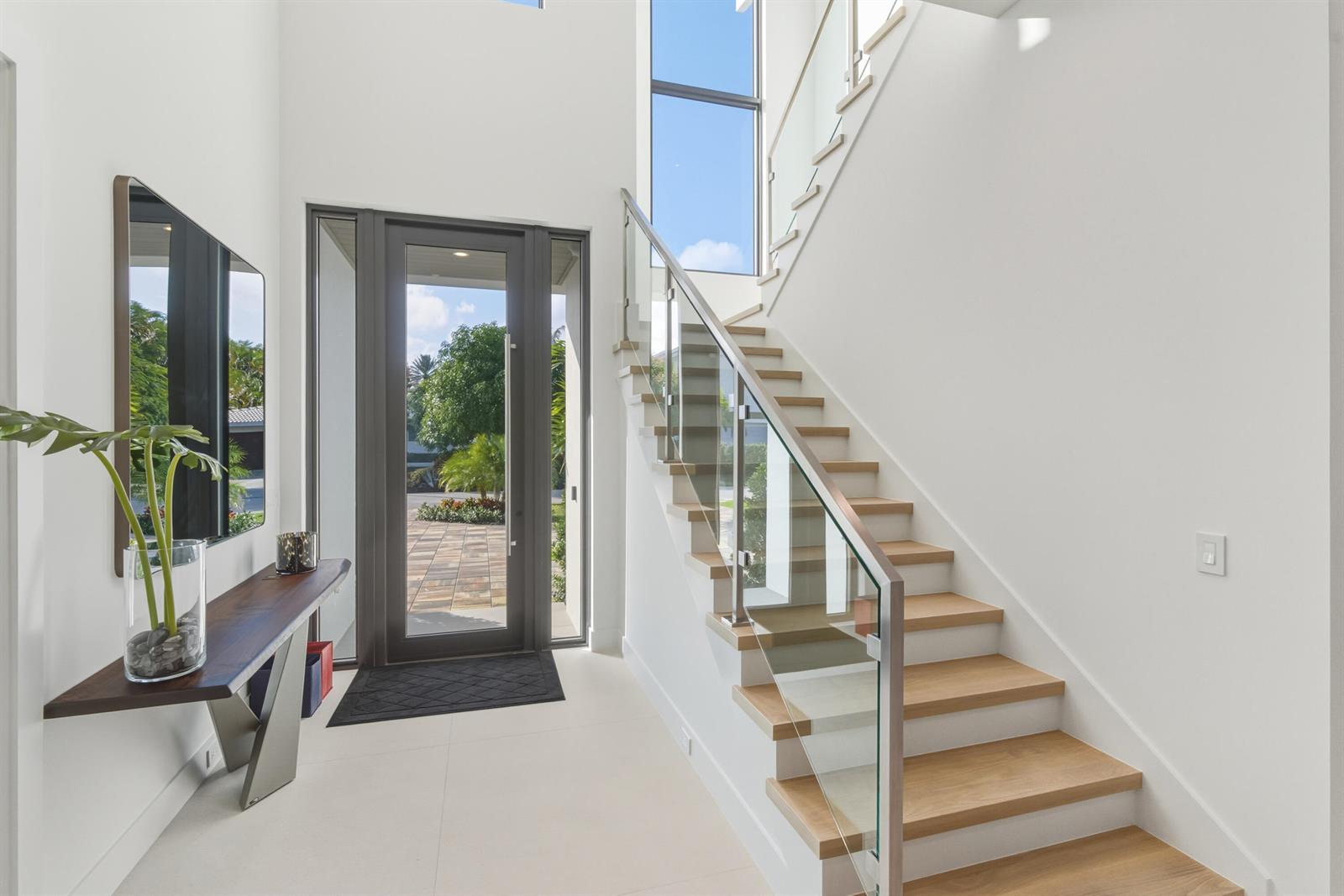 A modern entryway features a glass-paneled front door, a floating console table, and wooden stairs — 450 NE Spanish Court, Boca Raton FL 33432, BOCA RATON, FL