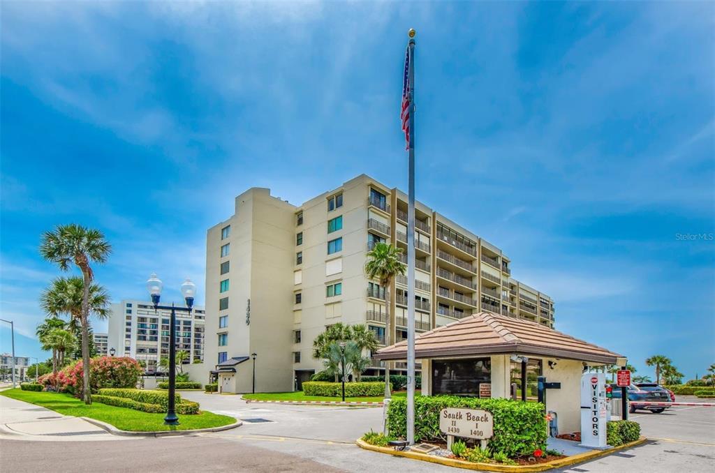 An exterior view of a multi-story apartment complex with a gated entrance and American flag — 1430 GULF BOULEVARD UNIT 612, CLEARWATER BEACH, FL