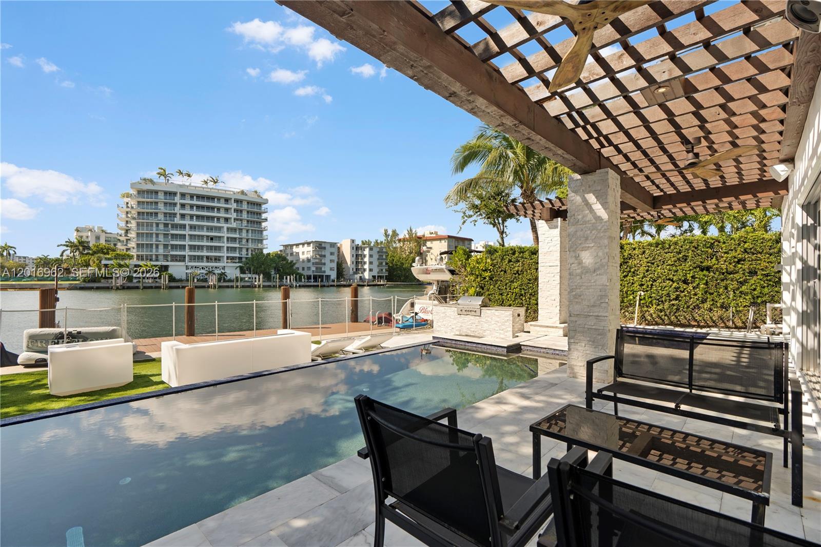 A patio features a pool, outdoor kitchen, and pergola overlooking a canal with docked boats — 9380 Bay Drive, Surfside FL 33154, SURFSIDE, FL