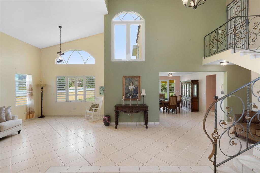 A bright, high-ceilinged living room with tile floors, a staircase, and a dining area beyond — CEIBA NORTE QUINTAS DE VALLE VERDE, JUNCOS, PR