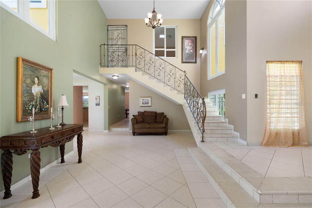 A spacious foyer features a tiled floor, a grand staircase with iron railings, and artwork — CEIBA NORTE QUINTAS DE VALLE VERDE, JUNCOS, PR