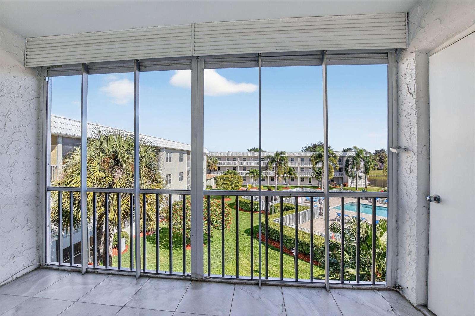 A tiled balcony with floor-to-ceiling windows overlooks a courtyard, swimming pool, and apartment buildings — 9872 Marina Boulevard # 1433, Boca Raton FL 33428, BOCA RATON, FL