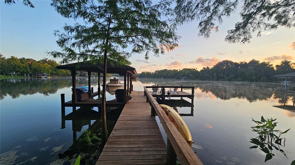 A wooden dock with a covered boat slip extends into a calm, tree-lined lake — 2021 WATERFORD ESTATES DRIVE, NEW SMYRNA BEACH, FL