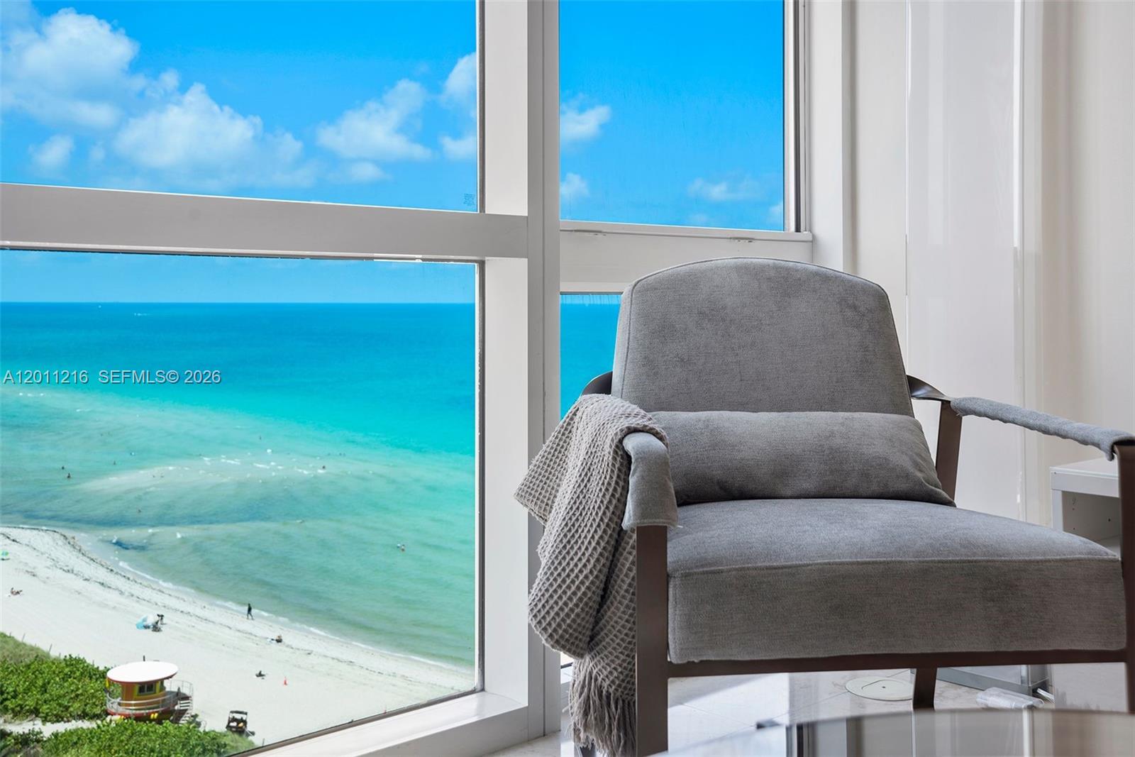A gray armchair sits next to a large window overlooking a beach and ocean — 6801 Collins Ave # 1203, Miami Beach FL 33141, MIAMI BEACH, FL