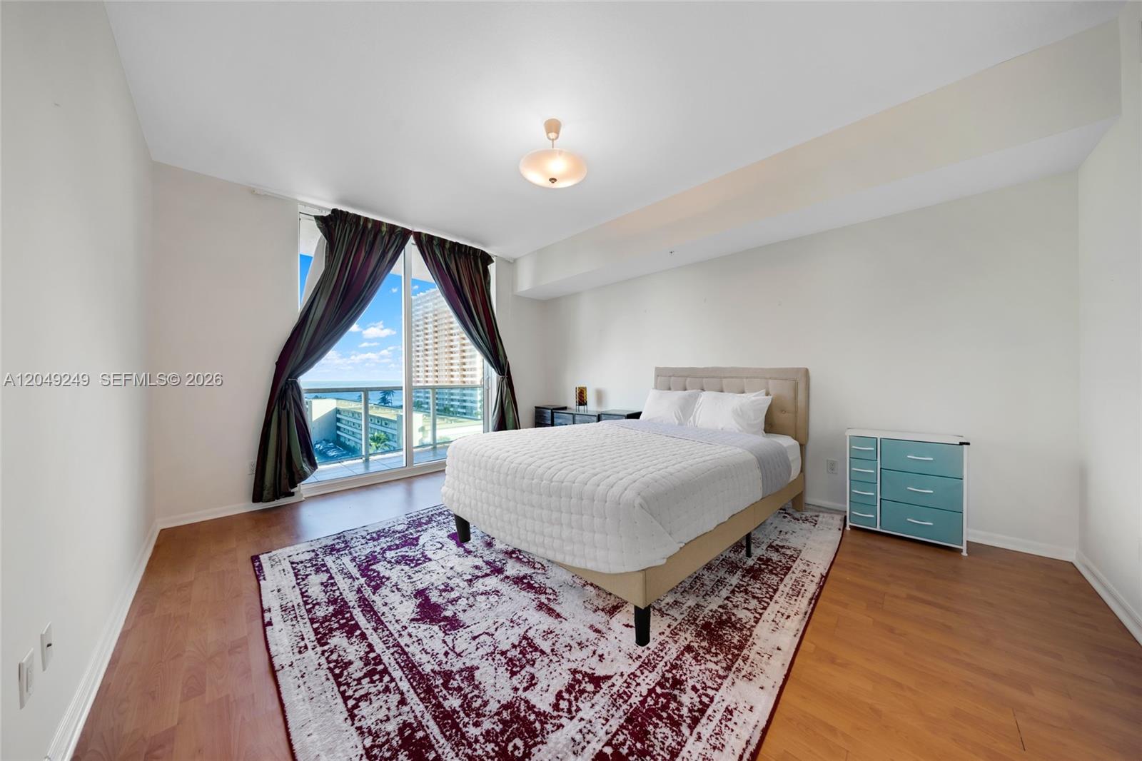 A bedroom features a bed, a patterned rug, a blue dresser, and a balcony view — 1945 S Ocean Dr # 714, Hallandale Beach FL 33009, HALLANDALE BEACH, FL