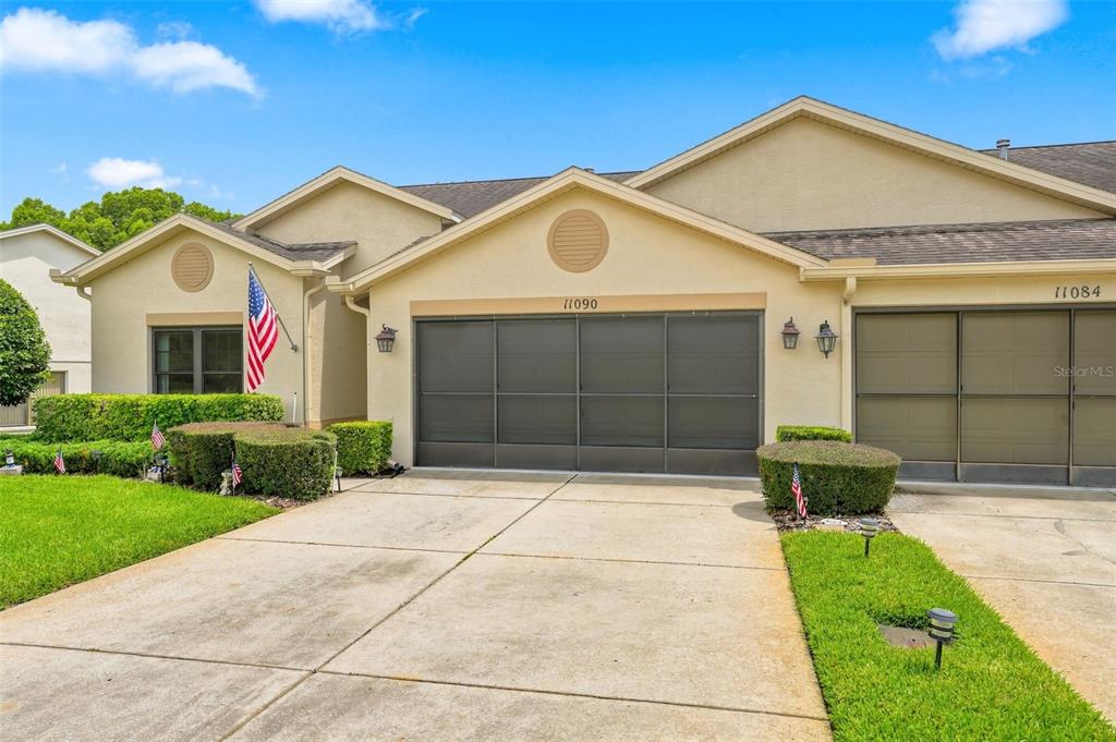 A beige duplex home features two garage doors, a concrete driveway, and manicured landscaping — 11090 CHERRYWOOD COURT, SPRING HILL, FL