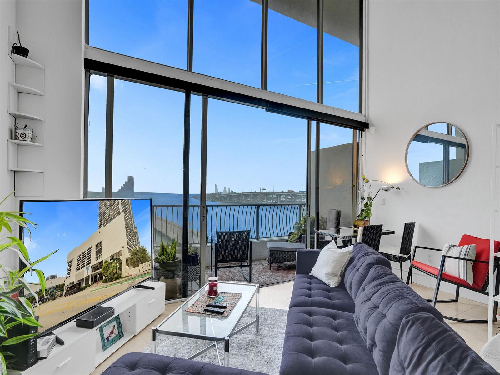 A living room with a tufted sofa, glass coffee table, and floor-to-ceiling windows overlooking water — 555 NE 15th Street, Miami FL 33132, MIAMI, FL