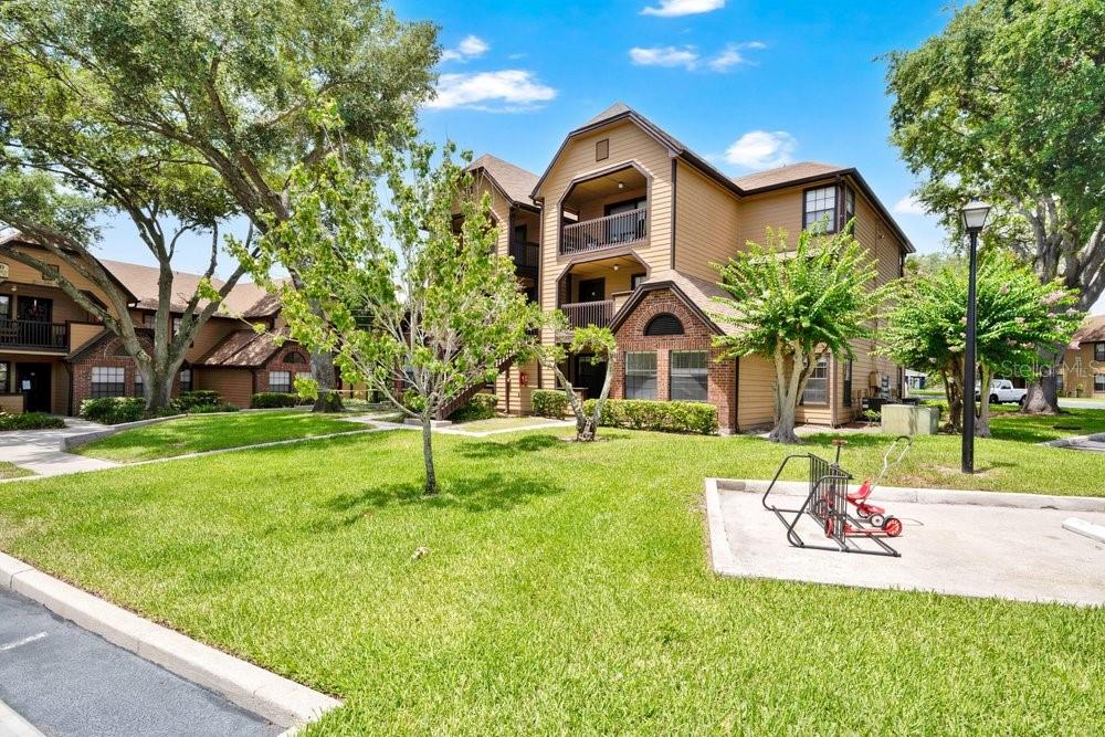 A three-story apartment building with tan siding stands behind a grassy lawn and bike rack — 360 LAKE ONTARIO COURT UNIT 104, ALTAMONTE SPRINGS, FL