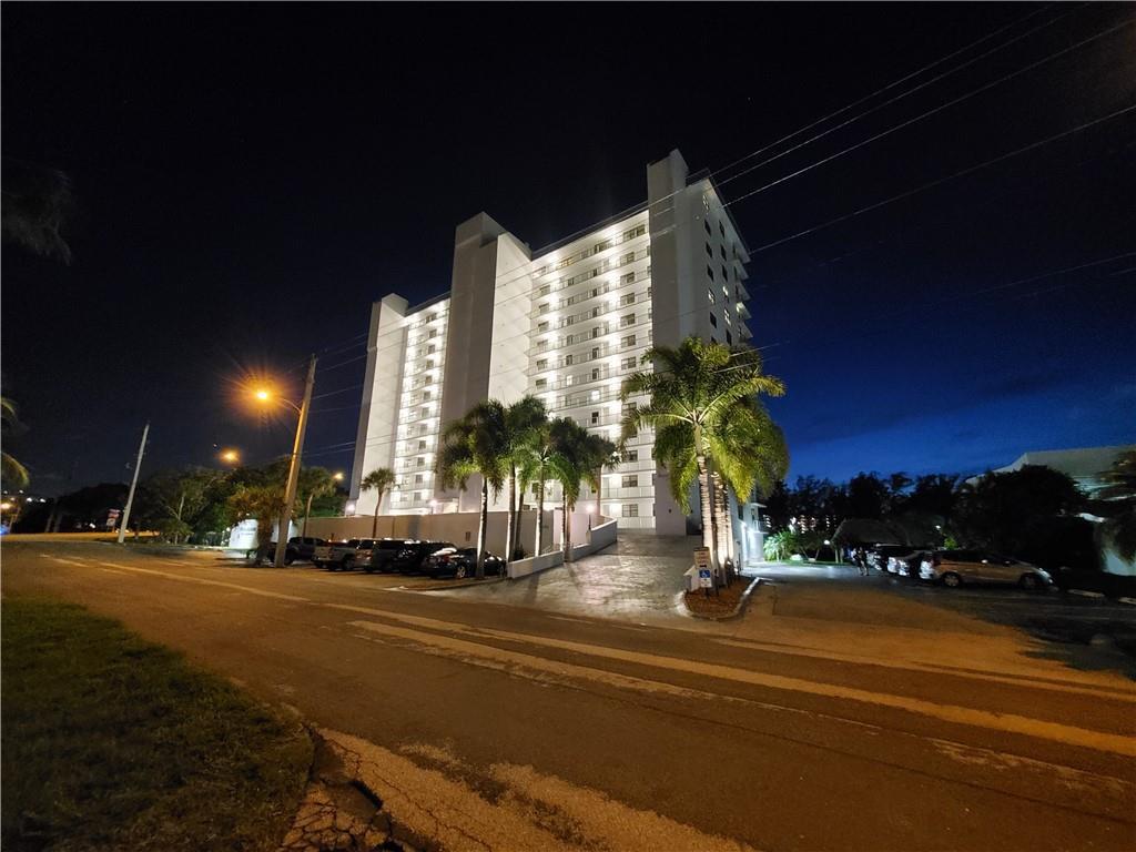 A tall, illuminated white apartment building stands at night next to a paved road — 1401 N Riverside Drive N # 807, Pompano Beach FL 33062, POMPANO BEACH, FL