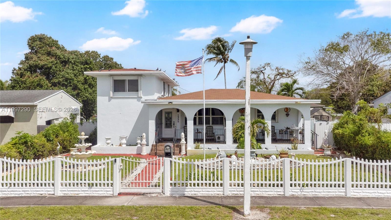 A white house with a covered porch and arched openings sits behind a white fence — 1335 NW 129th St, North Miami FL 33167, NORTH MIAMI, FL