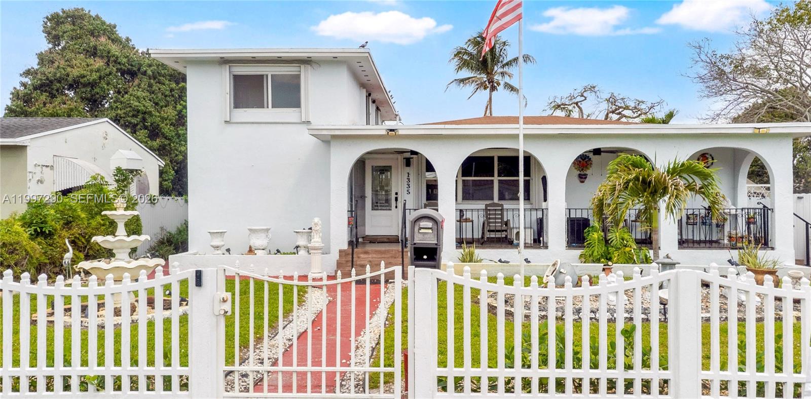A white two-story house features an arched front porch, a red walkway, and a fence — 1335 NW 129th St, North Miami FL 33167, NORTH MIAMI, FL