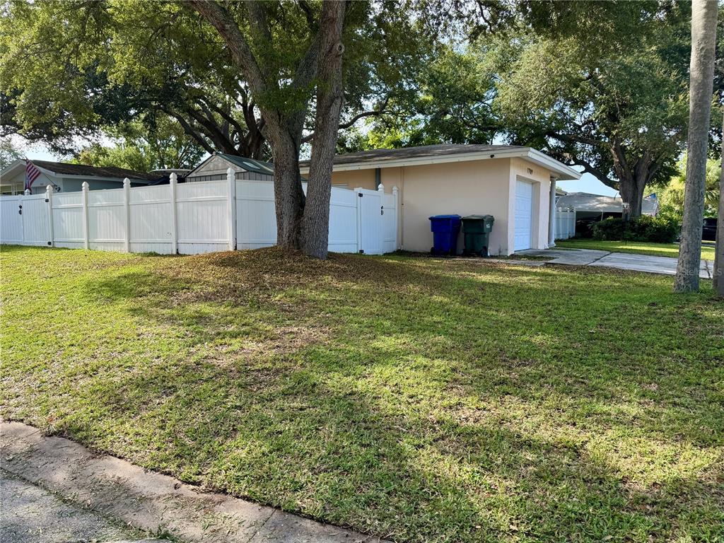 A beige house with a white fence and a garage sits behind a grassy yard — 1709 EATON DRIVE NE, CLEARWATER, FL