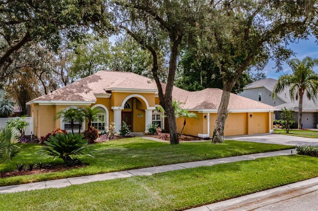A single-story yellow house with a three-car garage and arched entryway sits behind mature trees — 10131 PADDOCK OAKS DRIVE, RIVERVIEW, FL