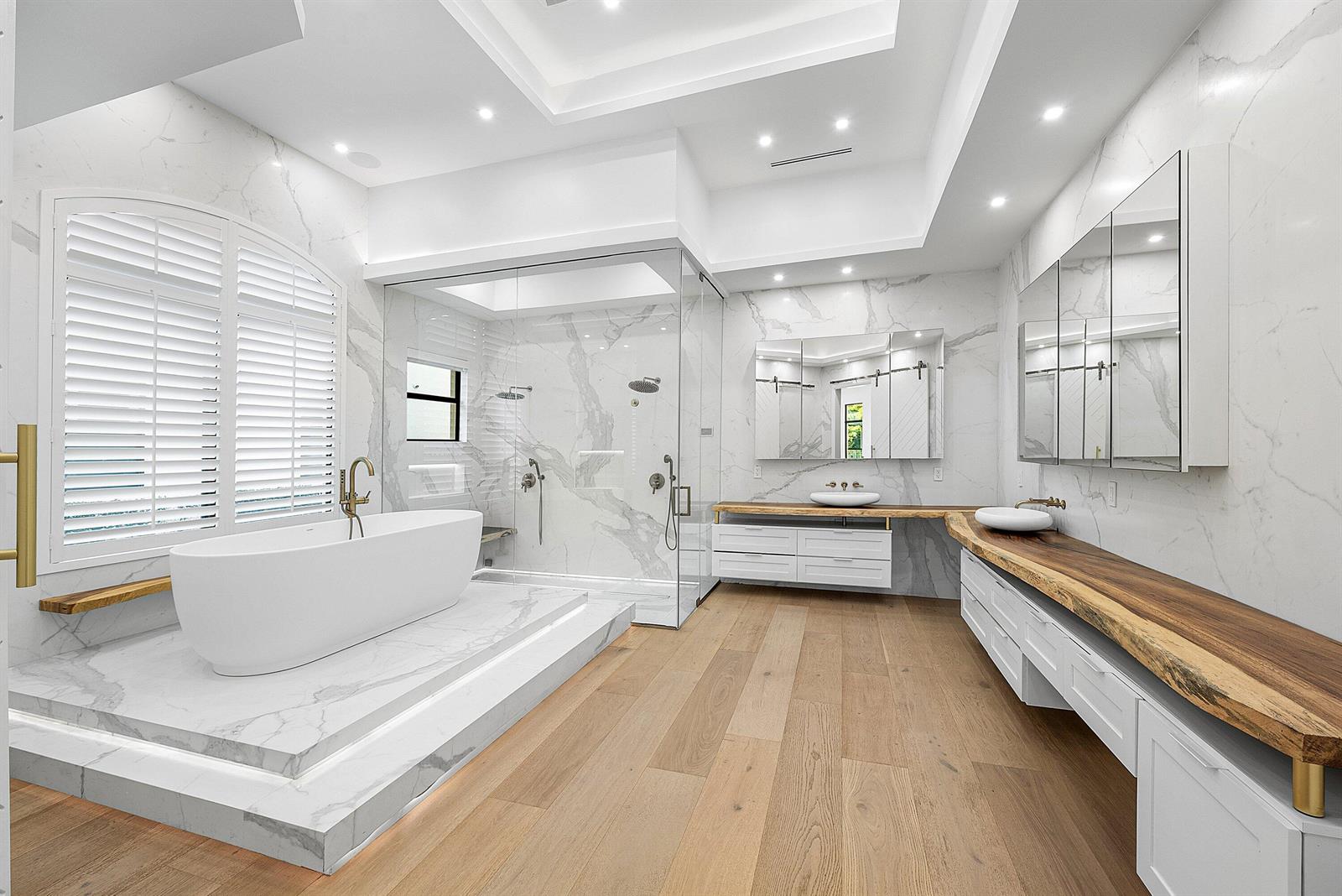 A modern bathroom features a marble-tiled soaking tub platform, glass shower, and live-edge wood vanities — 9599 Moritz Way, Delray Beach FL 33446, DELRAY BEACH, FL