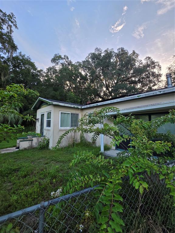 A single-story house exterior with a chain-link fence, overgrown vegetation, and large oak trees — 11647 SE 60TH AVENUE, BELLEVIEW, FL