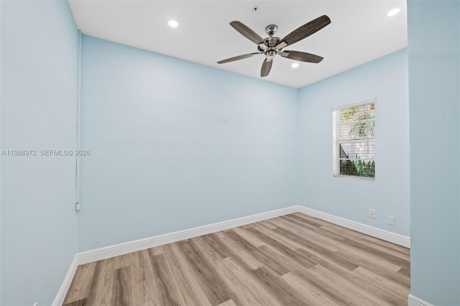 An empty room features light blue walls, wood-look flooring, a ceiling fan, and a window — 1633 NW 4th St, Boca Raton FL 33486, BOCA RATON, FL