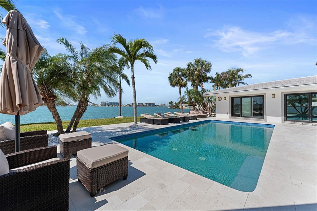A swimming pool sits on a stone patio overlooking a bay with palm trees — 11050 7TH STREET E, TREASURE ISLAND, FL