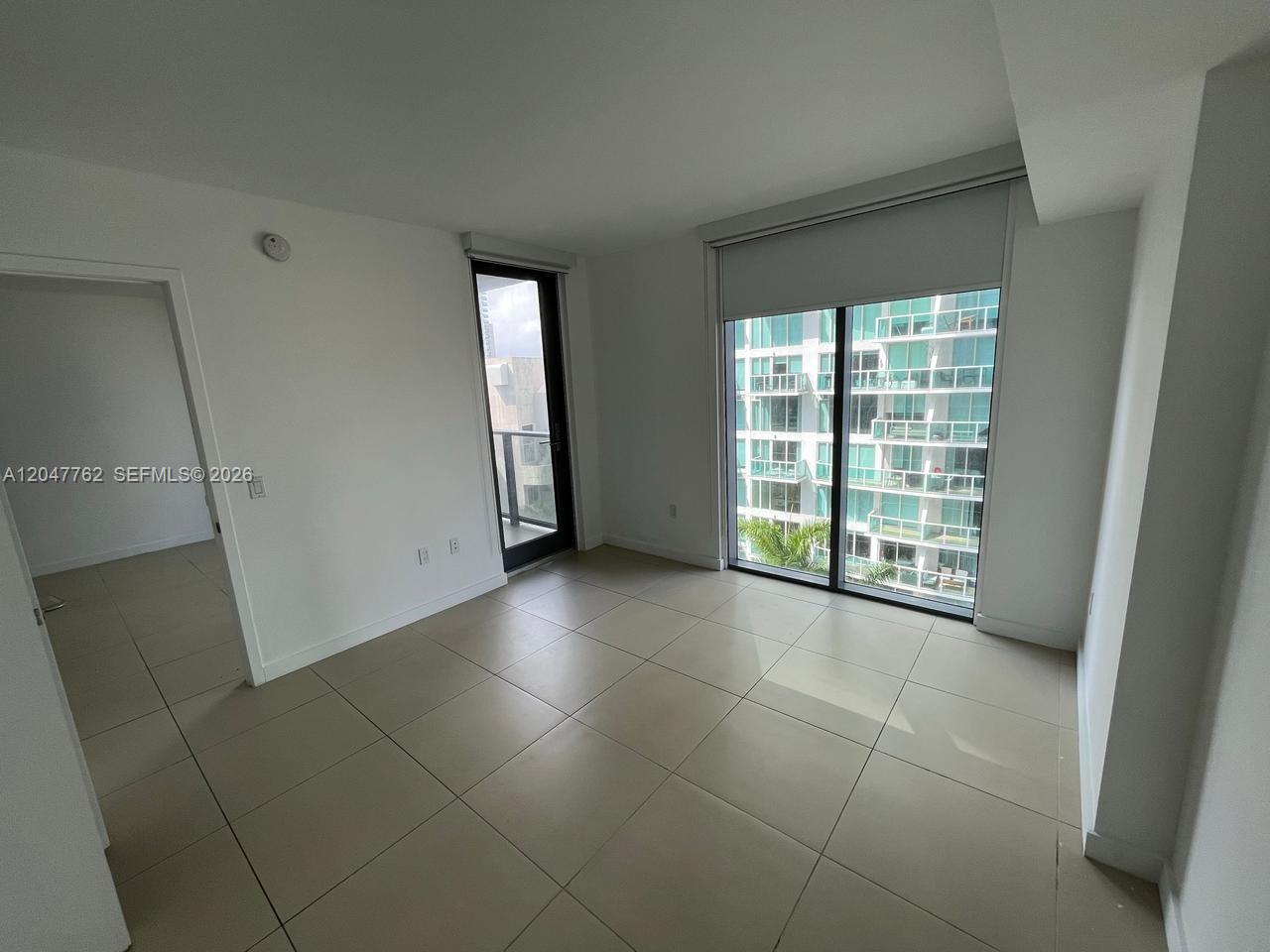An empty room features large light-colored floor tiles, white walls, and a glass balcony door — 31 SE 6th St # 1004, Miami FL 33131, MIAMI, FL