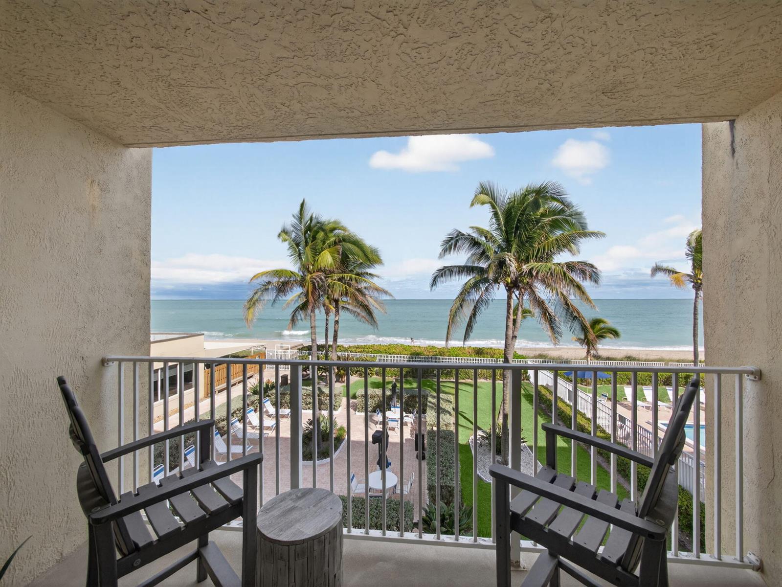 A balcony with two chairs and a small table overlooks a courtyard, palm trees, and ocean — 9800 S Ocean Drive # 302, Jensen Beach FL 34957, JENSEN BEACH, FL