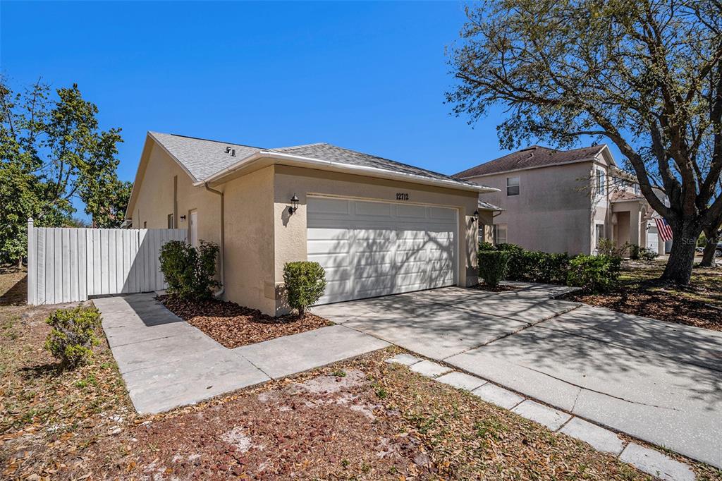 A beige house with a two-car garage and a white fence on a sunny day — 12712 LONGCREST DRIVE, RIVERVIEW, FL
