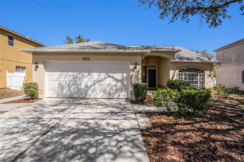 A single-story house with a two-car garage, beige stucco exterior, and a concrete driveway — 12712 LONGCREST DRIVE, RIVERVIEW, FL