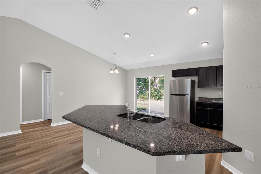 A kitchen features a granite-topped island, stainless steel refrigerator, dark cabinets, and wood-look flooring — 16 SPRING LOOP COURT, OCALA, FL
