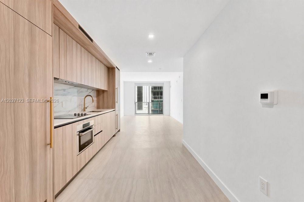 A modern kitchen features light wood cabinetry, stainless steel appliances, and white tile flooring — 77 SE 5th # 1711, Miami FL 33131, MIAMI, FL
