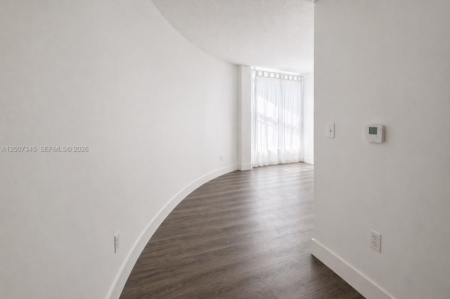 A curved white wall leads to a sunlit room with wood-look flooring and floor-to-ceiling curtains — 6580 INDIAN CREEK DR # 511, Miami Beach FL 33141, MIAMI BEACH, FL
