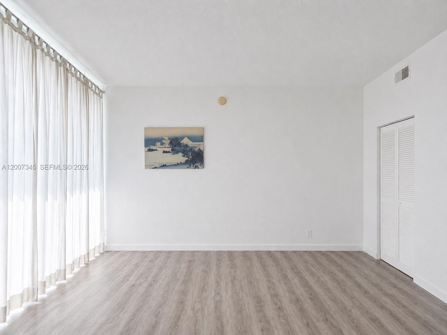 An empty room features white walls, light wood-look flooring, floor-to-ceiling curtains, and a closet — 6580 INDIAN CREEK DR # 511, Miami Beach FL 33141, MIAMI BEACH, FL