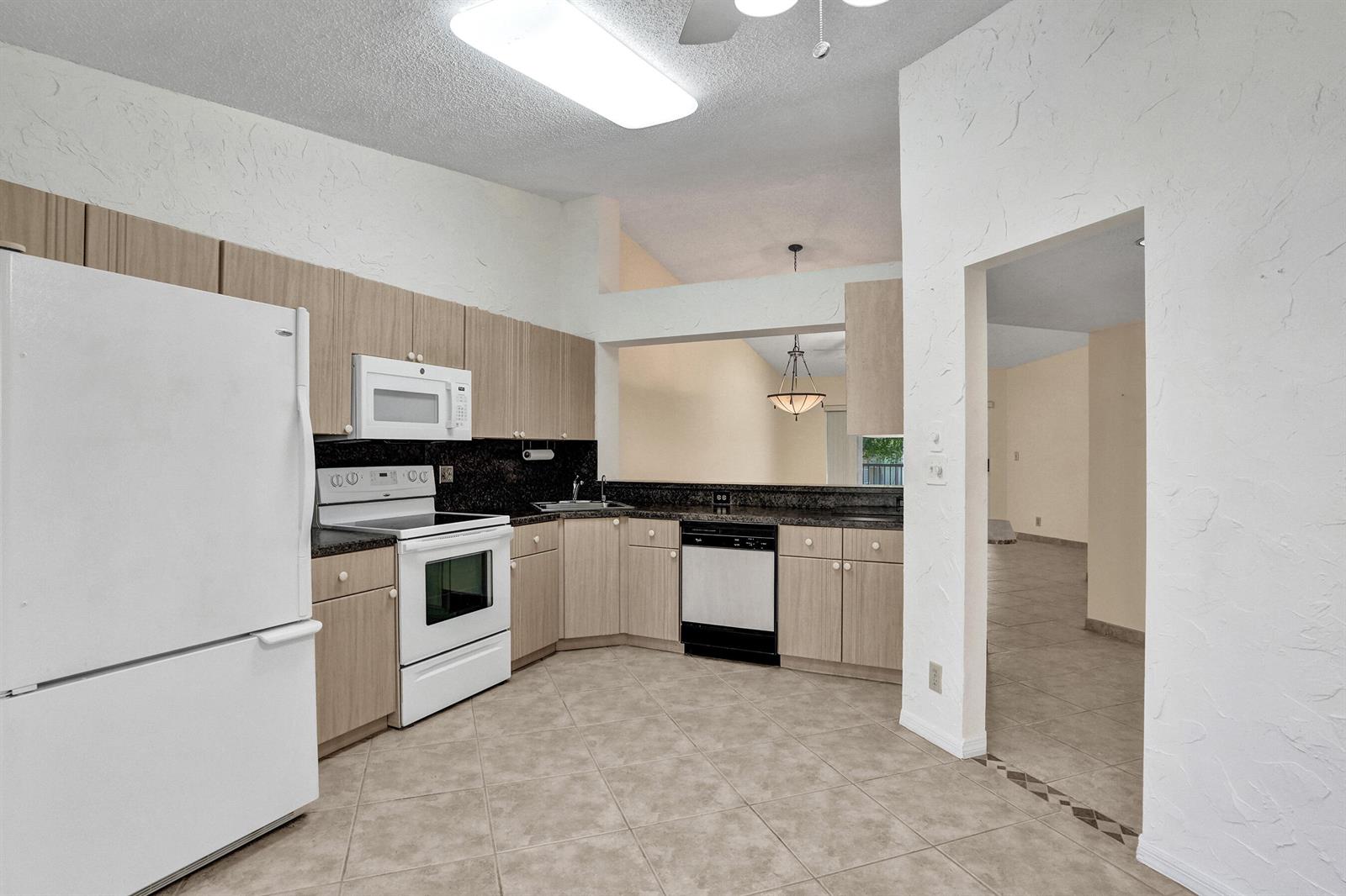 A kitchen features light wood cabinets, dark countertops, white appliances, and beige tile flooring — 7243 S Devon Drive, Tamarac FL 33321, TAMARAC, FL