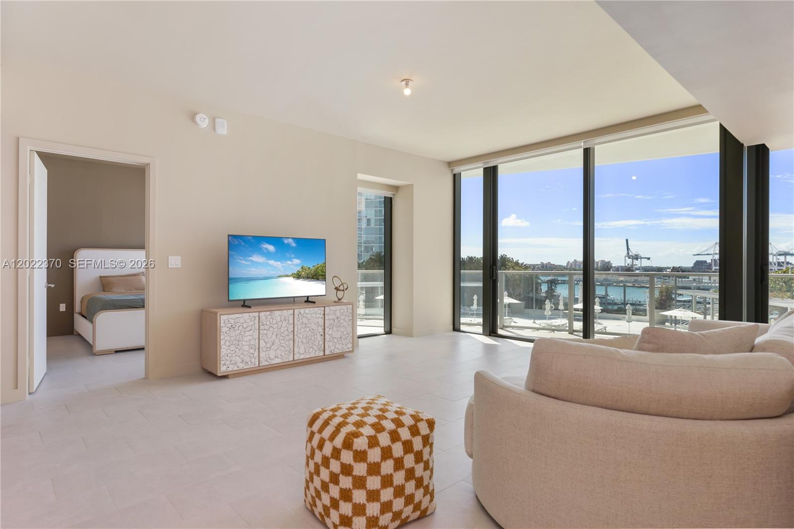 A living room features light tile floors, a television console, and floor-to-ceiling windows overlooking water — 500 Alton Rd # 802, Miami Beach FL 33139, MIAMI BEACH, FL