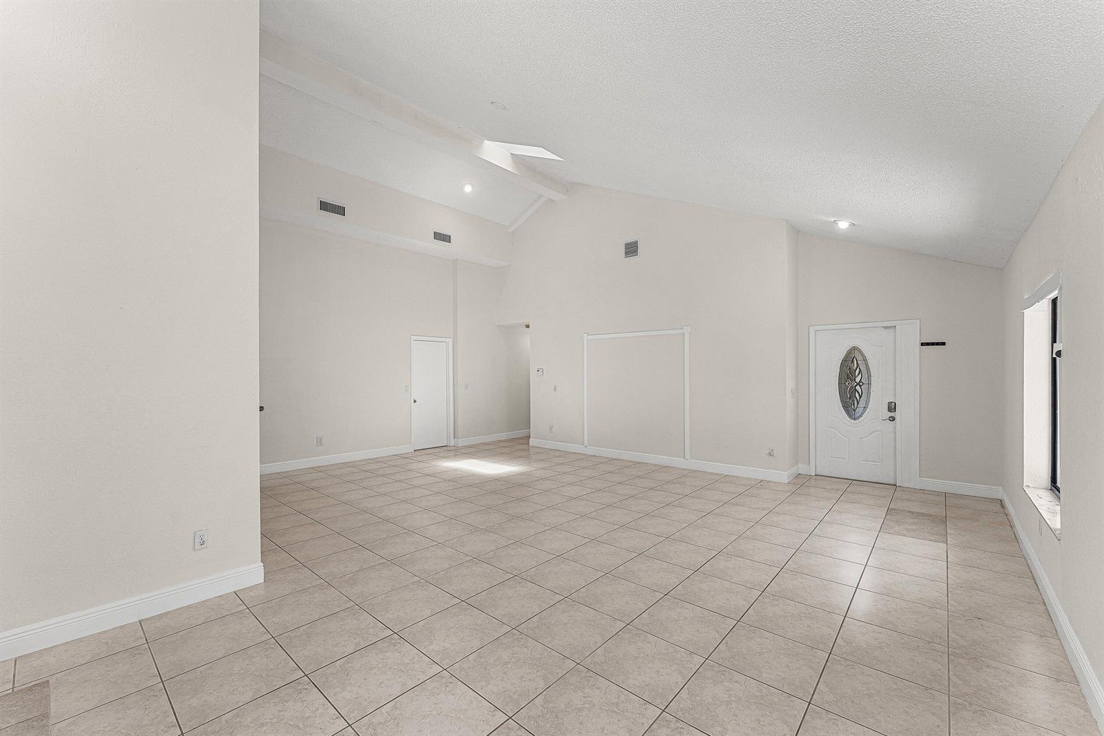 An empty room with vaulted ceilings, beige tile flooring, and a white front door — 7627 Sierra Drive W, Boca Raton FL 33433, BOCA RATON, FL