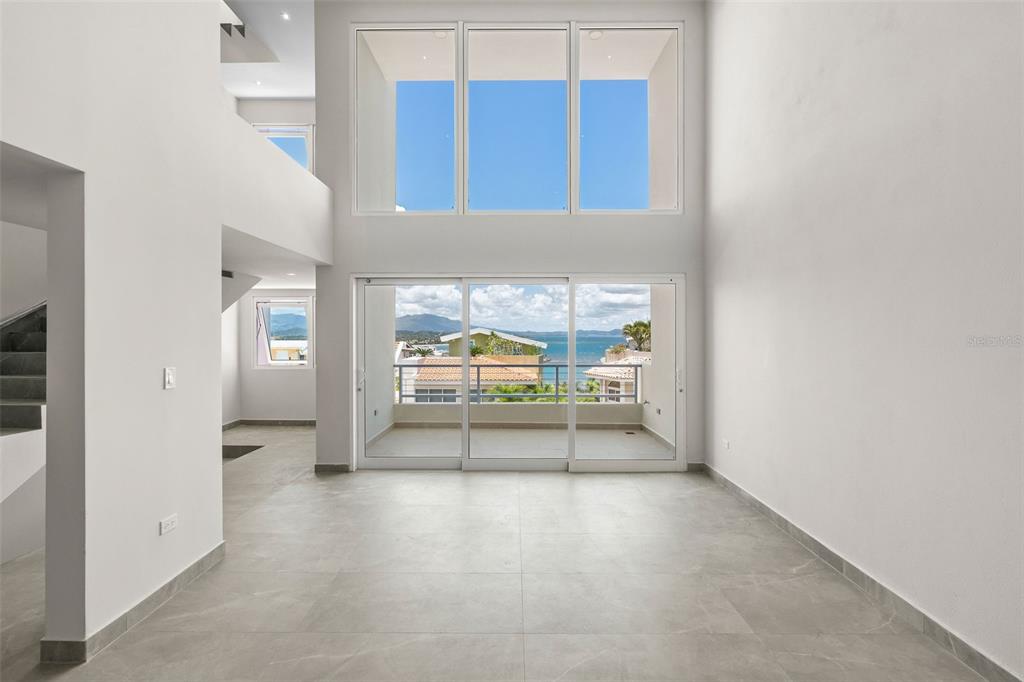 A bright, minimalist living room features double-height ceilings, large windows, and sliding glass doors overlooking the ocean — . SHELL CASTLE CLUB UNIT 46, HUMACAO, PR