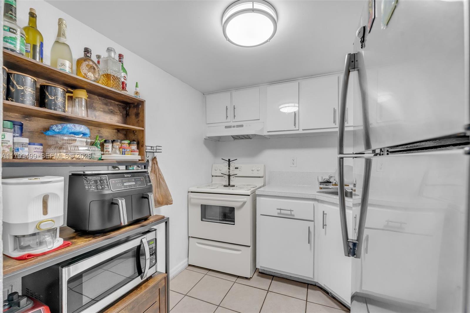 A small kitchen features white cabinets, white appliances, a tiled floor, and open wooden shelving — 4550 NW 9th St # 106E, Miami FL 33126, MIAMI, FL