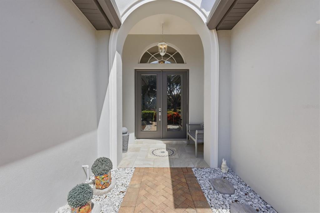 A covered entryway features a brick path, white gravel, double glass doors, and an arched ceiling — 7346 WINDEMERE LANE, UNIVERSITY PARK, FL