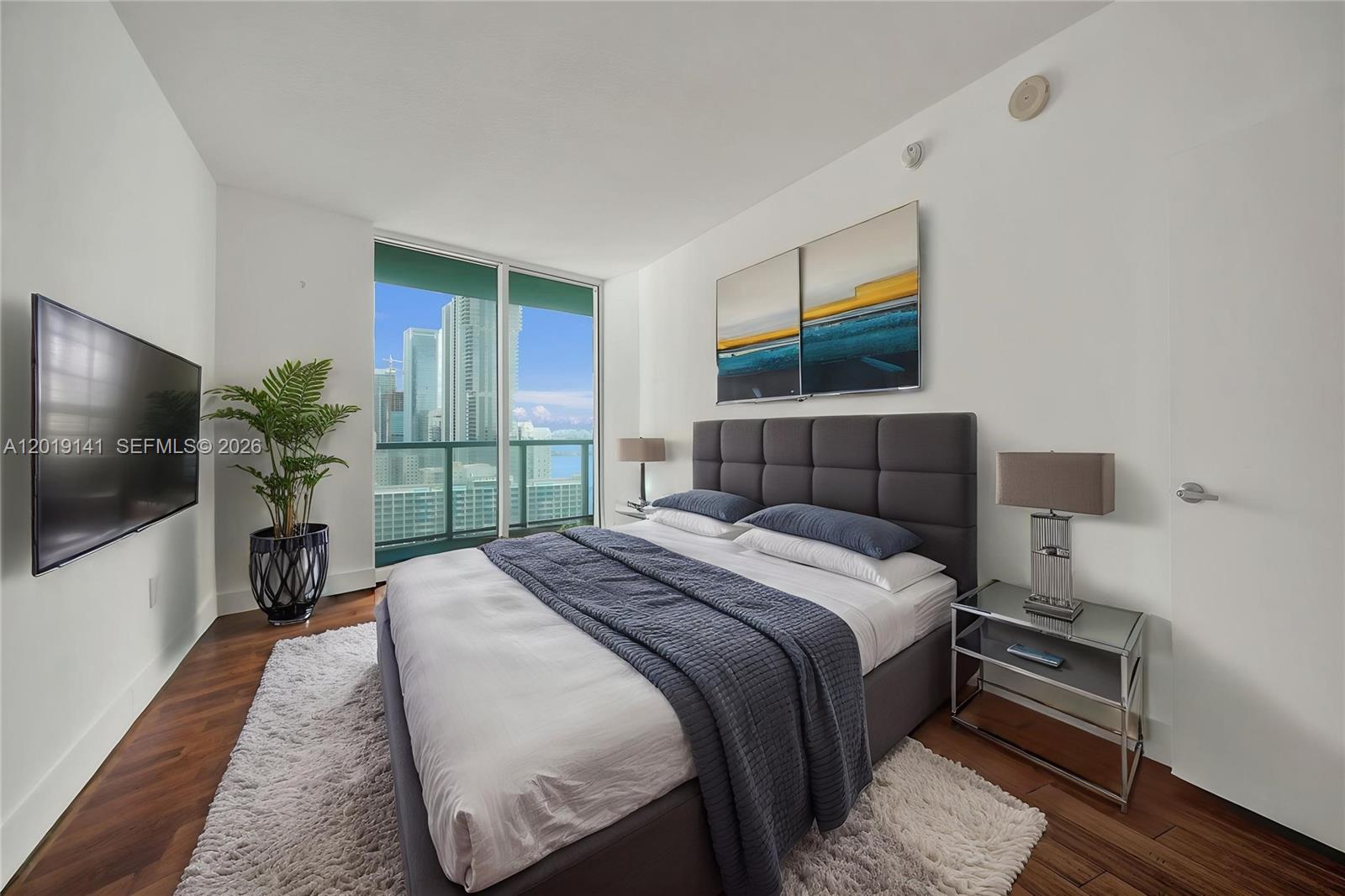A modern bedroom features a gray upholstered bed, mirrored nightstands, and a city skyline view — 1900 N Bayshore Dr # 2204, Miami FL 33132, MIAMI, FL