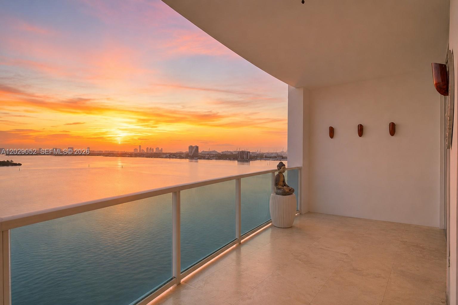 A balcony with a glass railing overlooks a sunset over the water and city skyline — 725 NE 22nd St # 11E, Miami FL 33137, MIAMI, FL
