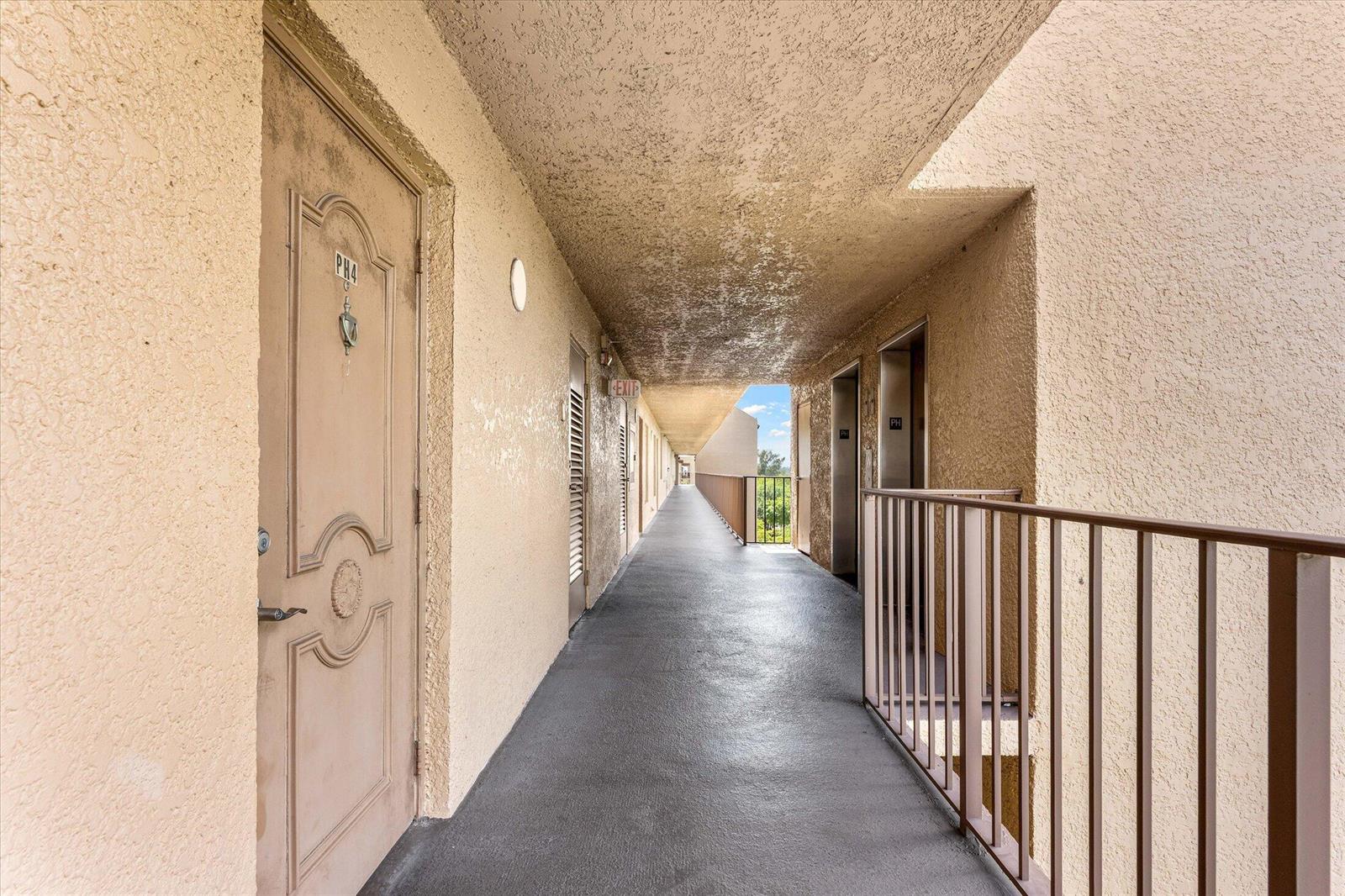 An outdoor apartment hallway with beige stucco walls, a gray floor, and a metal railing — 4570 NW 18th Avenue # 704, Deerfield Beach FL 33064, DEERFIELD BEACH, FL