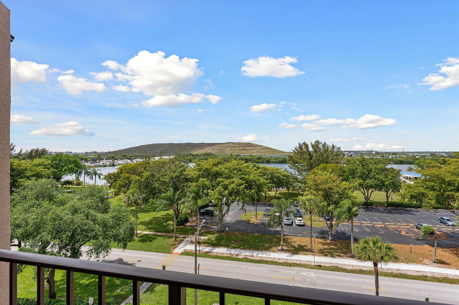 A balcony view overlooking a parking lot, green trees, a body of water, and a hill — 4570 NW 18th Avenue # 704, Deerfield Beach FL 33064, DEERFIELD BEACH, FL