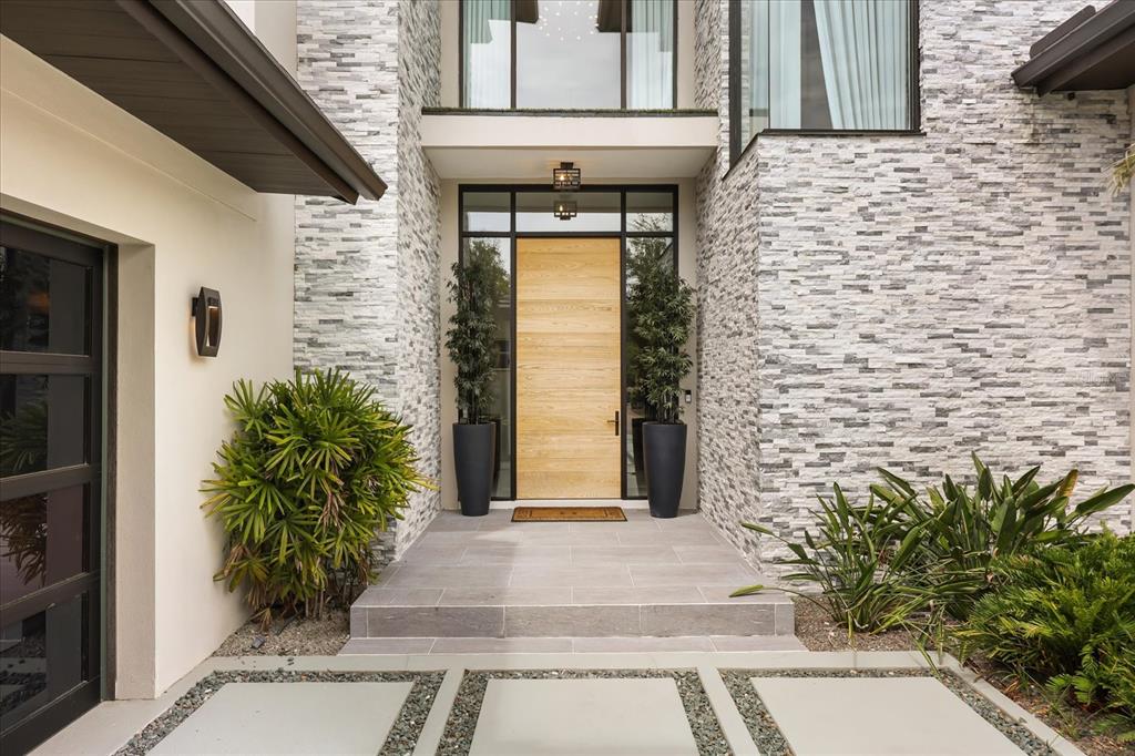 A modern home entrance features a light wood door, stone veneer walls, and potted plants — 10348 KENSINGTON SHORE DRIVE, ORLANDO, FL