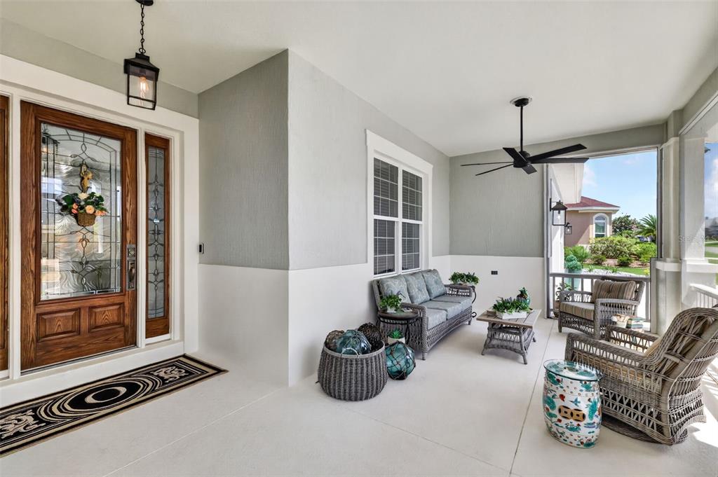 A covered porch features a wooden front door, wicker furniture, and a ceiling fan — 3468 MCDANIEL PLACE, THE VILLAGES, FL