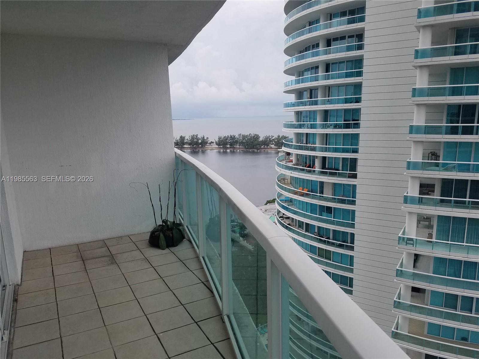 A balcony with tiled flooring overlooks a body of water and a neighboring high-rise building — 2101 Brickell Ave # 1911, Miami FL 33129, MIAMI, FL