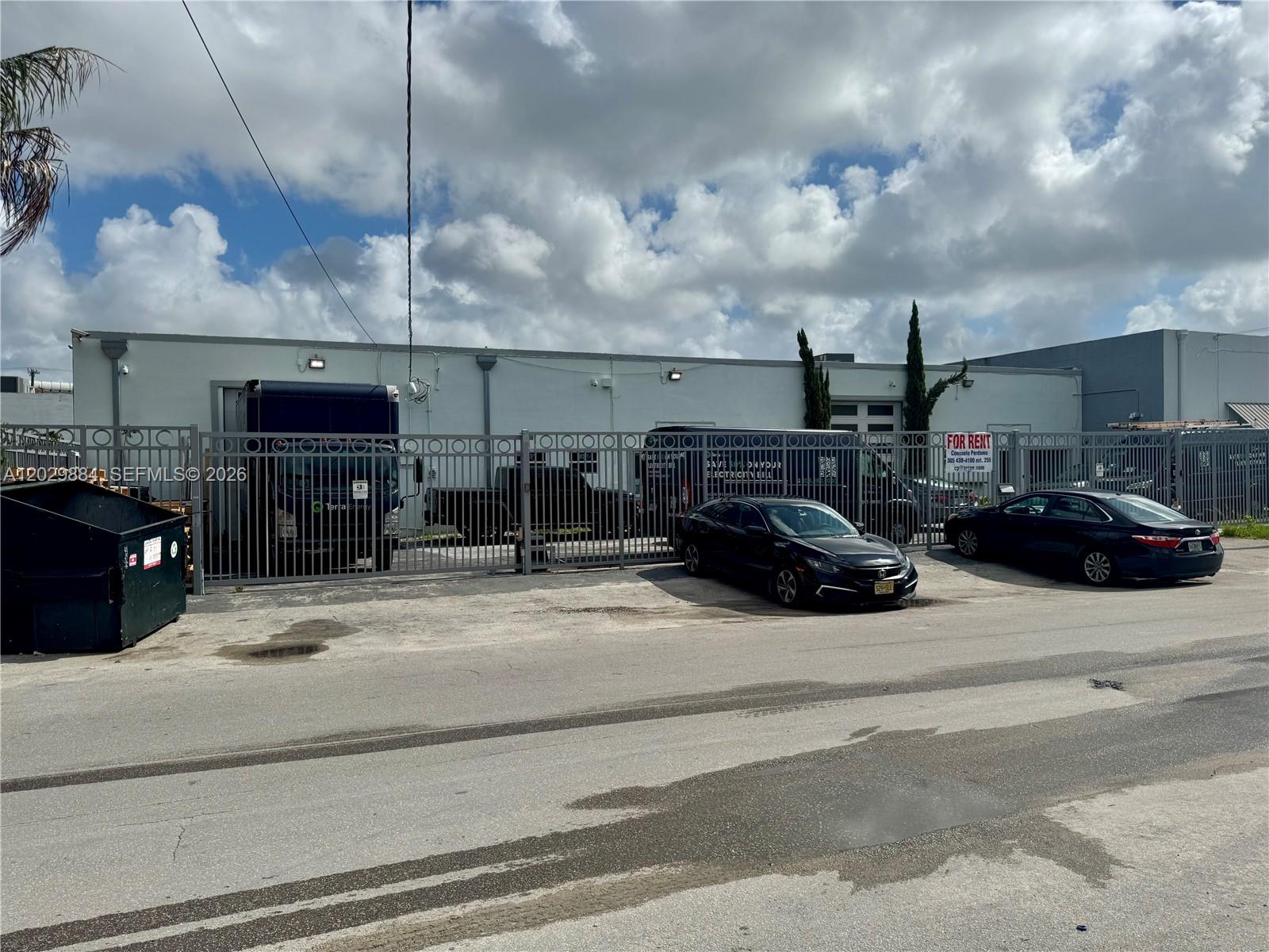 A fenced industrial building exterior features a parking area with several vehicles and a dumpster — 3241 NW 38th St, Miami FL 33142, MIAMI, FL