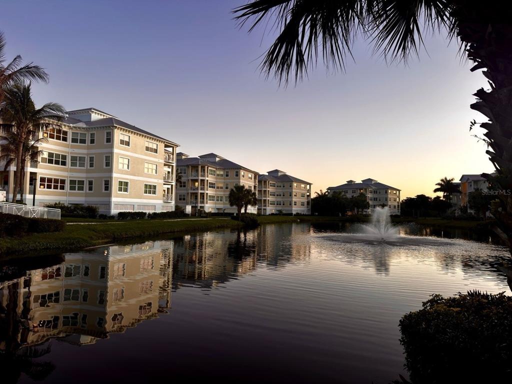 A multi-story apartment complex sits beside a pond with a fountain at sunset — 3418 79TH STREET CIRCLE W UNIT 101, BRADENTON, FL