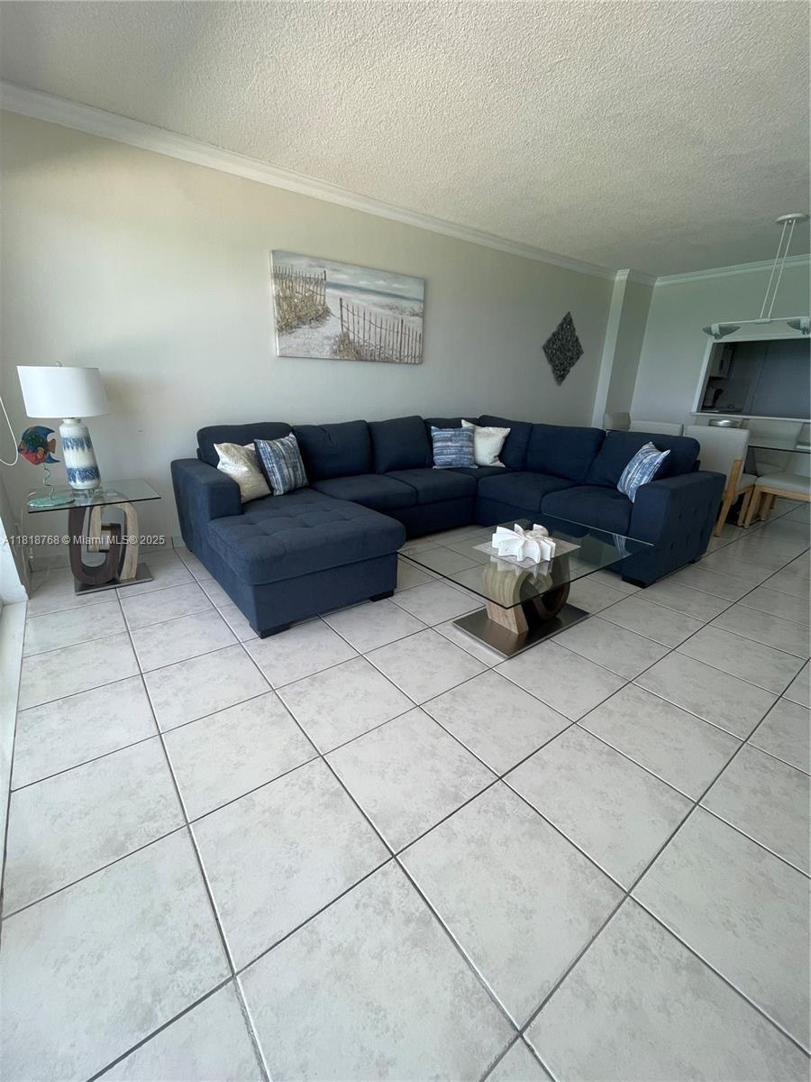 A living room features a large blue sectional sofa, a glass coffee table, and tile flooring — 5601 Collins # 1210, Miami Beach FL 33140, MIAMI BEACH, FL