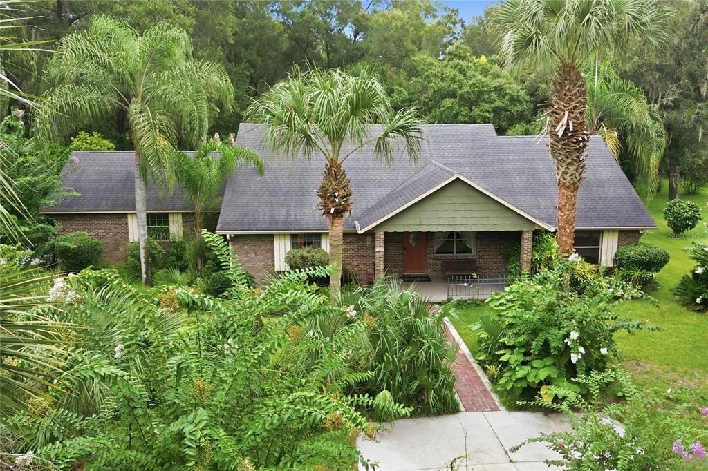 A brick ranch-style house with a covered porch, surrounded by lush landscaping and palm trees — 1925 LADY BUG LANE, DELAND, FL