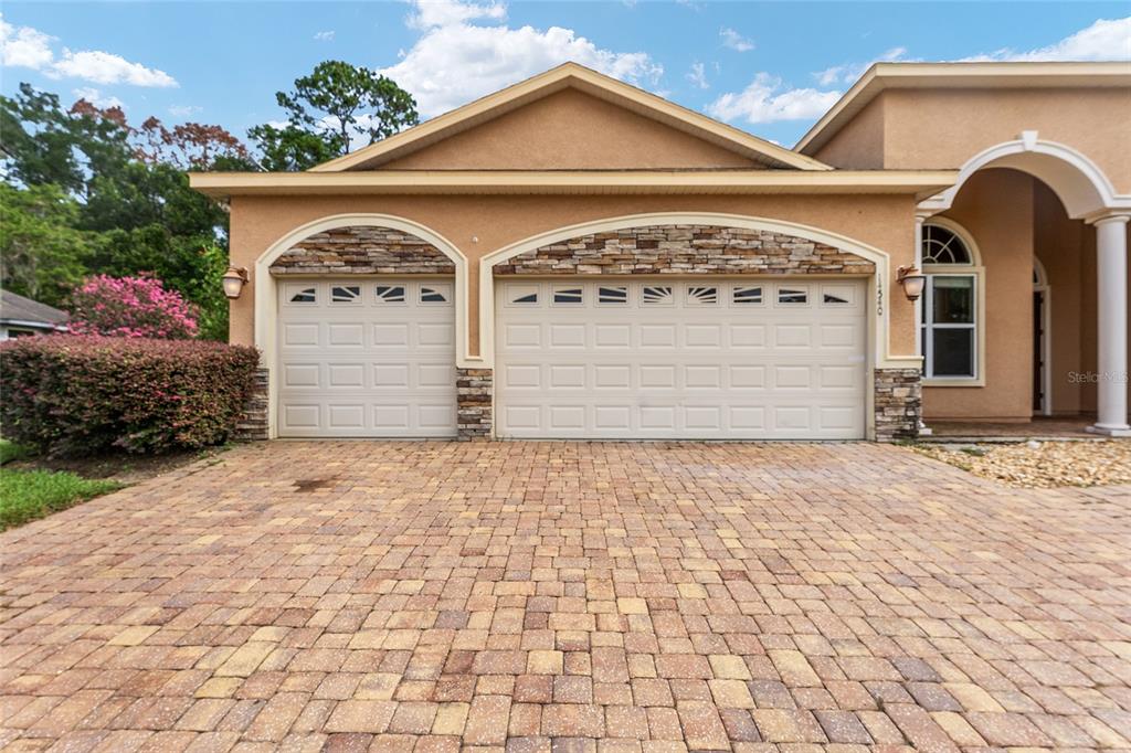 A tan house exterior features a three-car garage with stone accents and a paver driveway — 14540 NW 27TH PLACE, NEWBERRY, FL