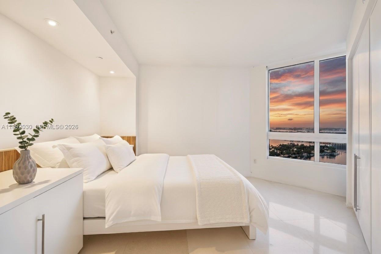 A modern bedroom features a white bed, tiled floors, and a large window overlooking water — 6365 Collins Ave # 2901, Miami Beach FL 33141, MIAMI BEACH, FL